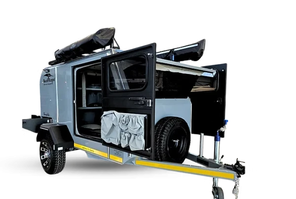 Side view of a grey Bush Lapa Shongololo off-road caravan with side doors fully open, showing internal storage shelves, canvas storage pouches attached to the door, a spare wheel mounted near the hitch, and a rolled-up rooftop awning.