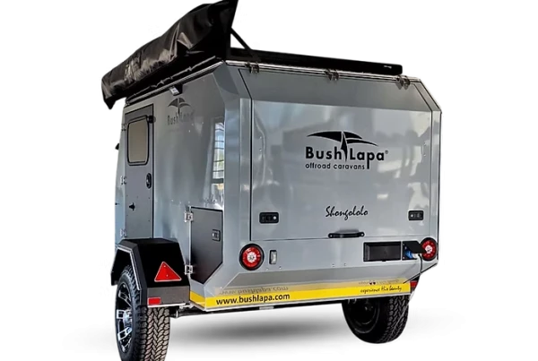 Three-quarter rear view of a grey Bush Lapa Shongololo off-road caravan showing its rugged metallic body, off-road tire, rear taillights, electrical plug inlet, and black awning bag mounted on top.