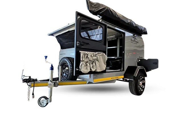 Front three-quarter view of a grey Bush Lapa Shongololo caravan showcasing the drawbar and jockey wheel, front-mounted spare tire, side access door with canvas organizer pockets open to reveal the interior sleeping/storage area, and a top-mounted awning.